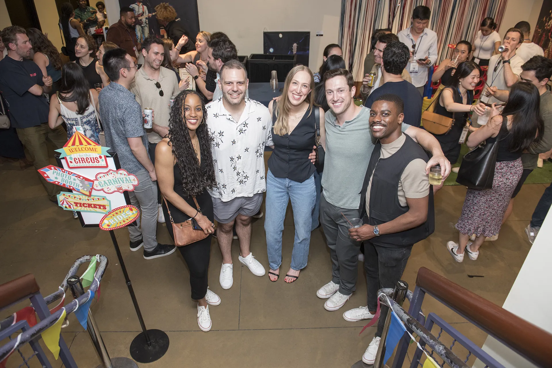 A group of Class of 2015 graduates smile and gather around a carnival-themed sign reading “Photo Booth” and “Tickets” at their 10-year Reunion event