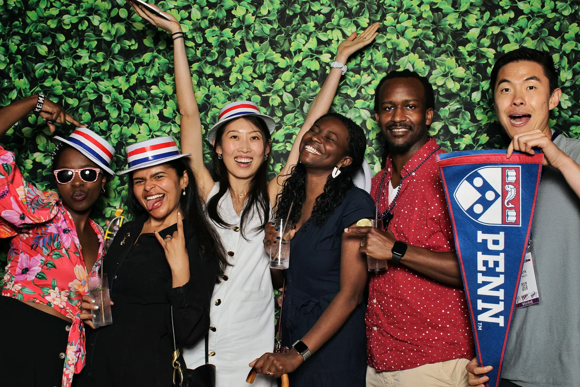 Six Penn alumni from the Class of 2015 cheer in a photo booth wearing red, white, and blue hats and holding drinks and a Penn pennant in front of a leafy green backdrop