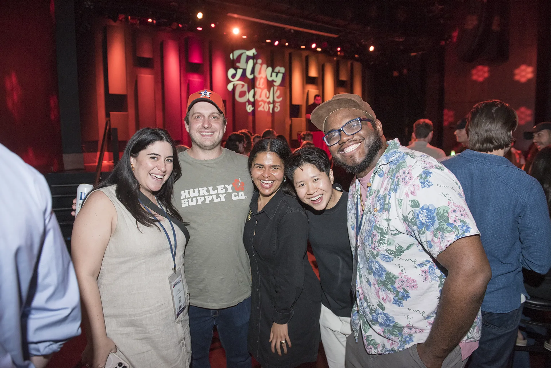 Smiling Penn alumni from the Class of 2015 posing together in front of a stage lit with pink and red lights during their 10th Reunion “Fling It Back” event