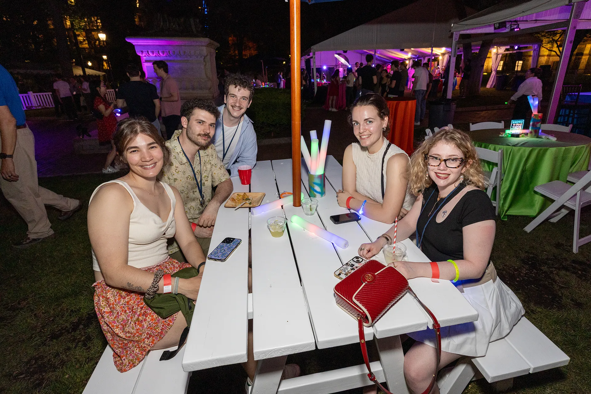 Group of Penn Class of 2020 alumni sitting at a white picnic table with glowing light sticks at the 5th Reunion night under a lit event tent
