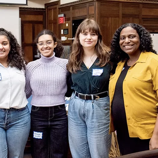 A group of women standing together in an indoor setting with wooden paneling and chairs in the background.