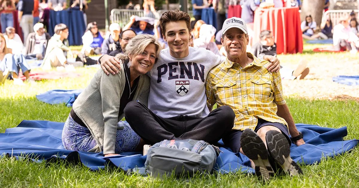 Student in a Penn sweatshirt poses with parents on a sunny day as families gather for an outdoor event on College Green.