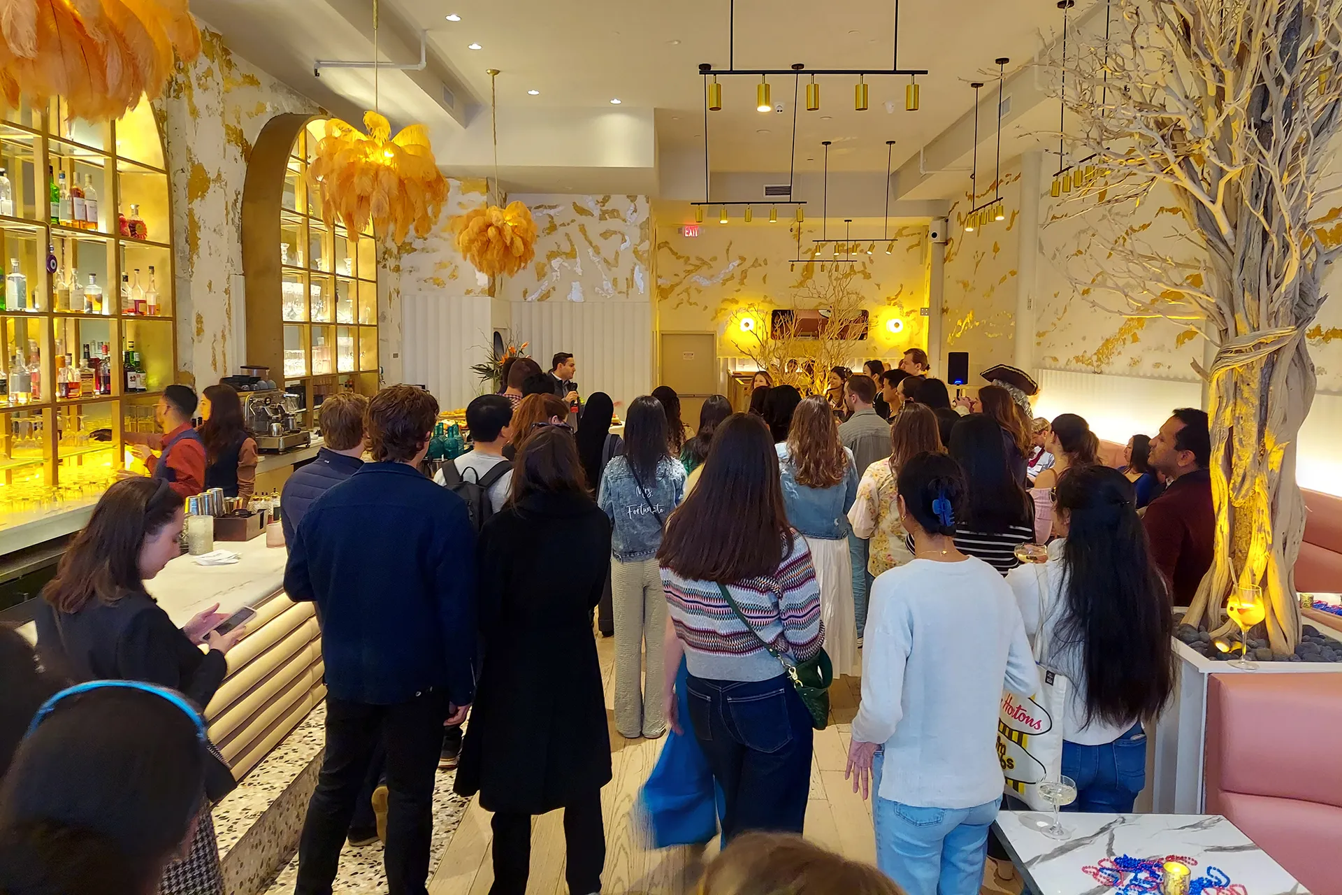A stylish, gold-accented bar in New York City filled with alumni standing and listening as a host delivers a toast near the back wall.