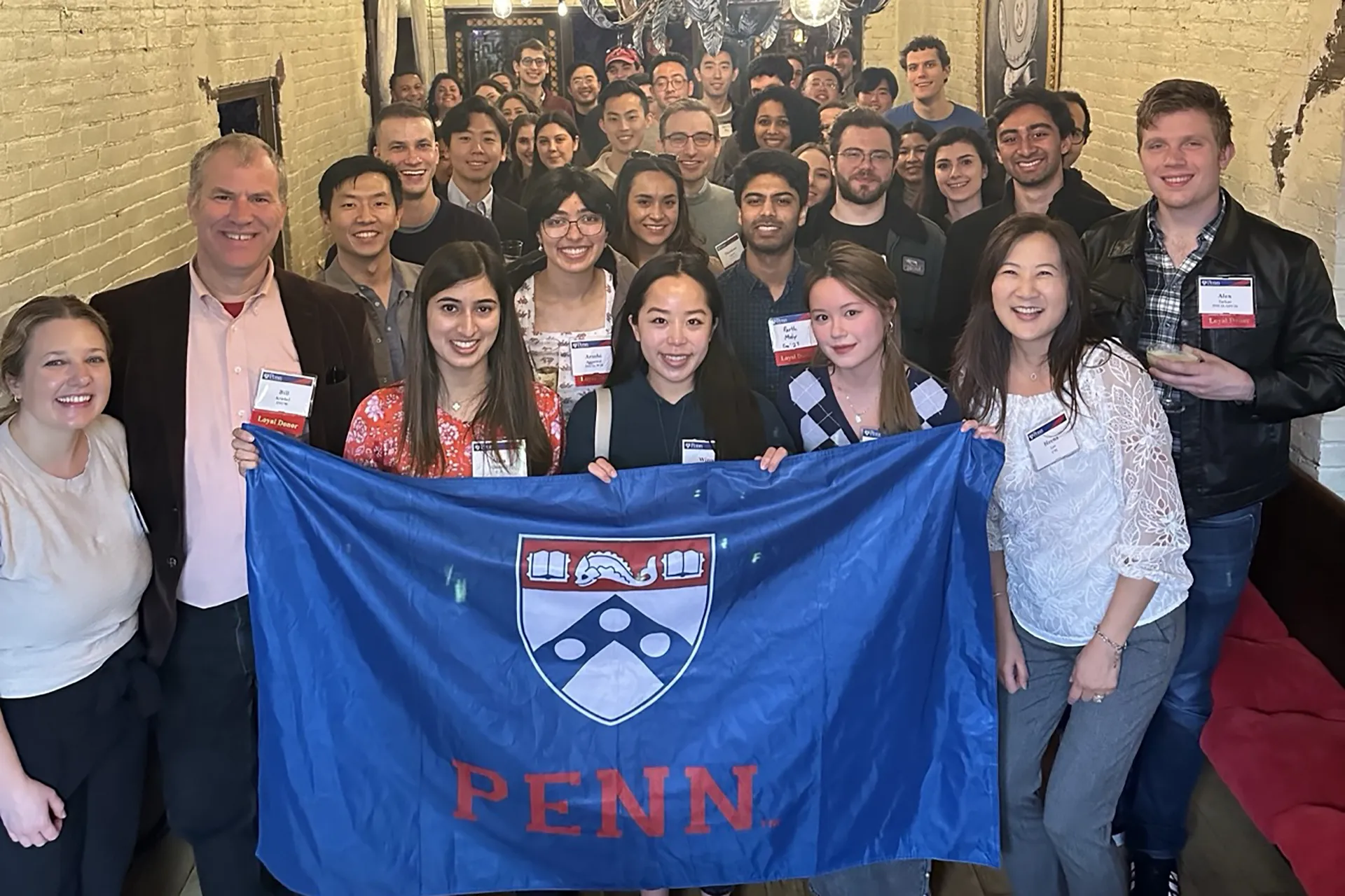 A large group of Penn alumni holding a blue “PENN” banner and smiling together at the 2024 First Toast on the Road event in Boston.