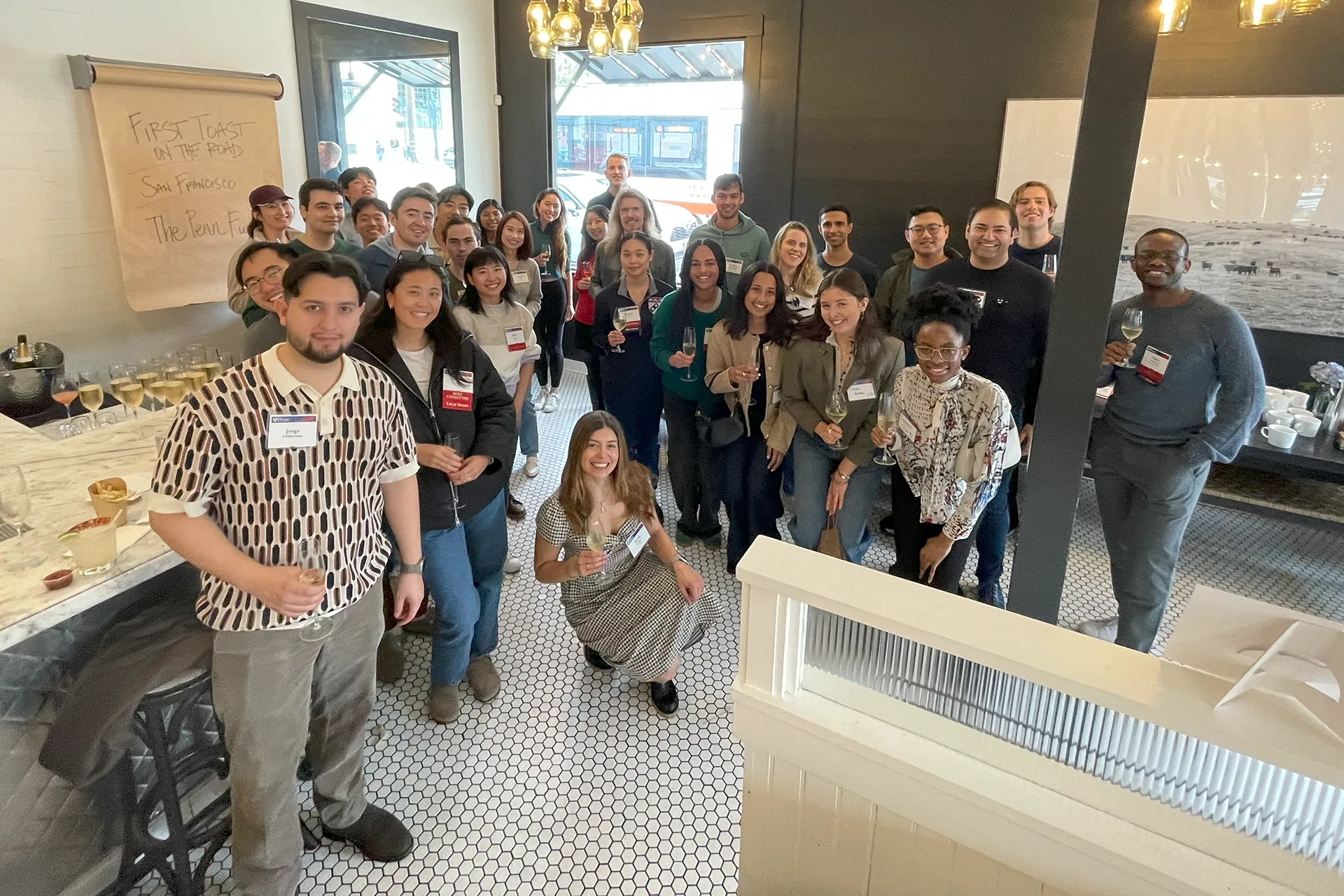 Smiling attendees raise their glasses in a sunlit room with black-trimmed windows and a flip-chart poster marking the First Toast on the Road event in San Francisco.
