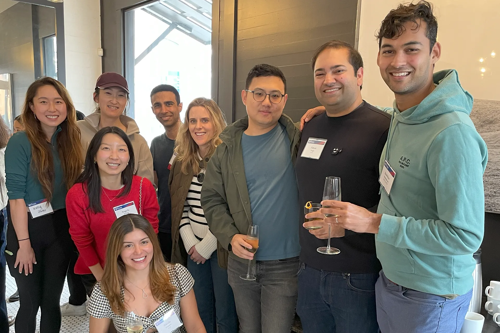 Alumni guests cluster under natural light by a windowed entrance, grinning and holding wine flutes at the SF First Toast gathering.