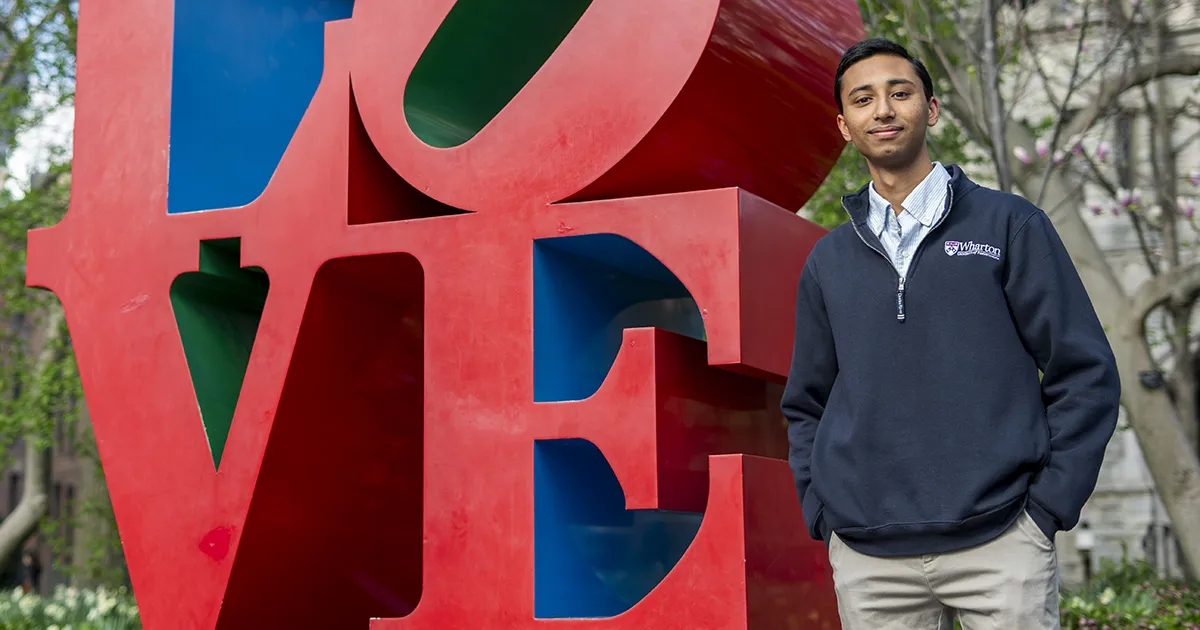Penn student in Wharton gear stands beside the iconic LOVE sculpture on campus during spring, representing Giving Red and Blue Day support for undergraduate education.