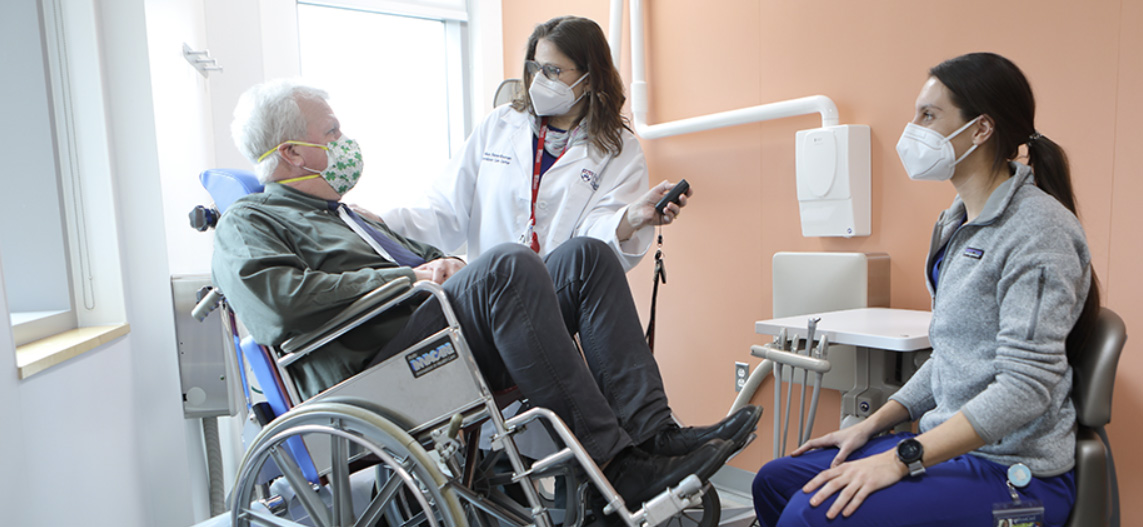 Two dental professionals speak with a patient who sits in a wheelchair.