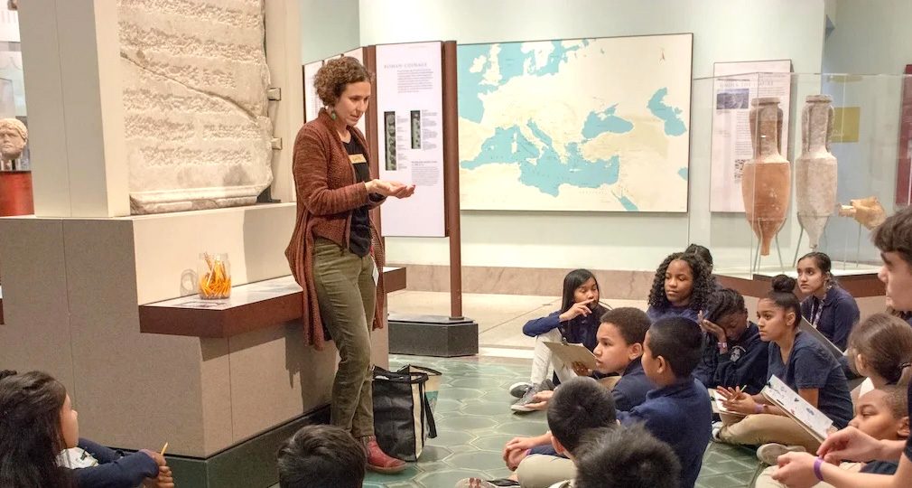 A museum educator speaks in a gallery with a class of students during their field trip to the Penn Museum.