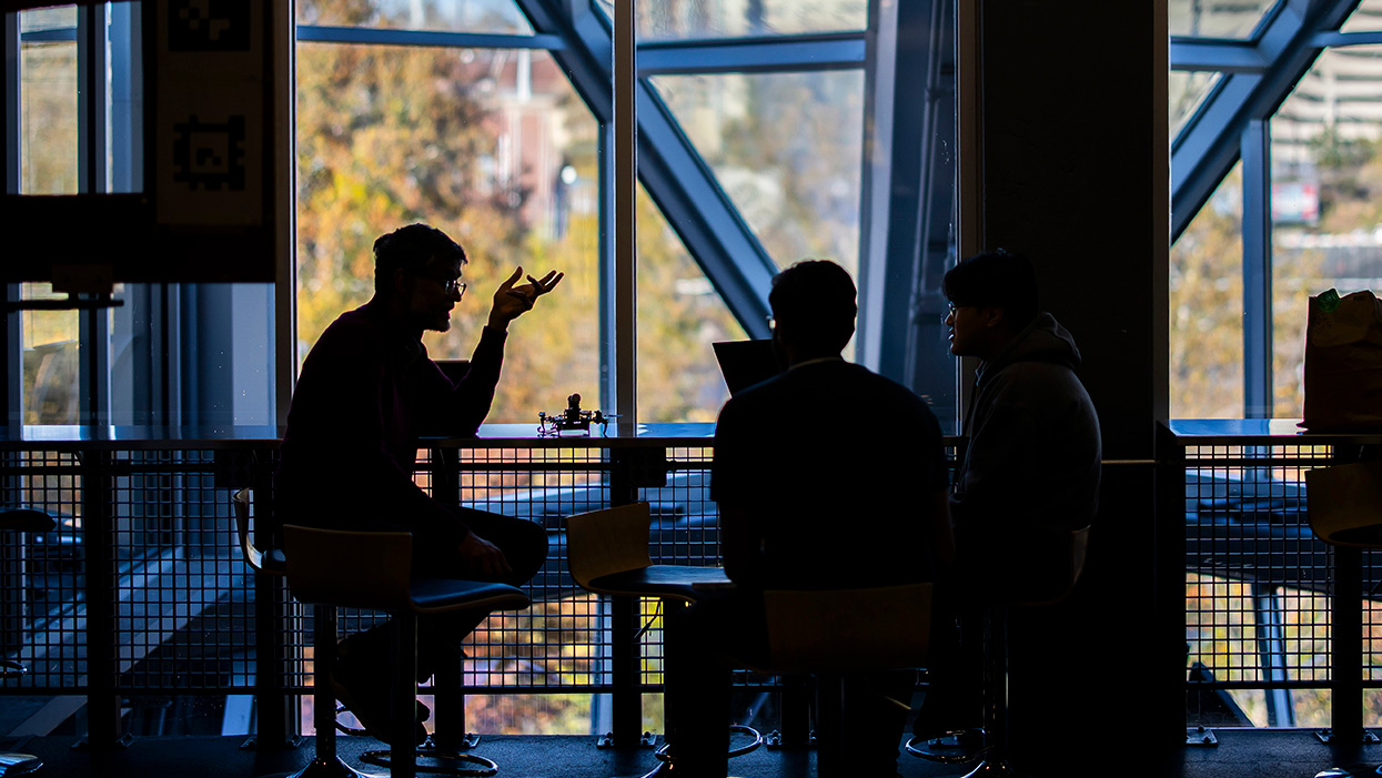 A group of engineering students, shown in silhouette against a windowed background, sit and speak at a table.