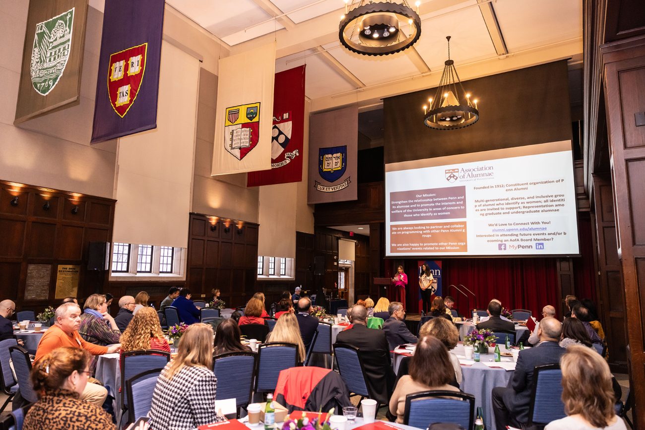 A formal gathering of diverse alumni seated at round tables in the Hall of Flags at Houston Hall on the University of Pennsylvania campus. Two speakers, one standing at the podium, are presenting to the attentive audience.