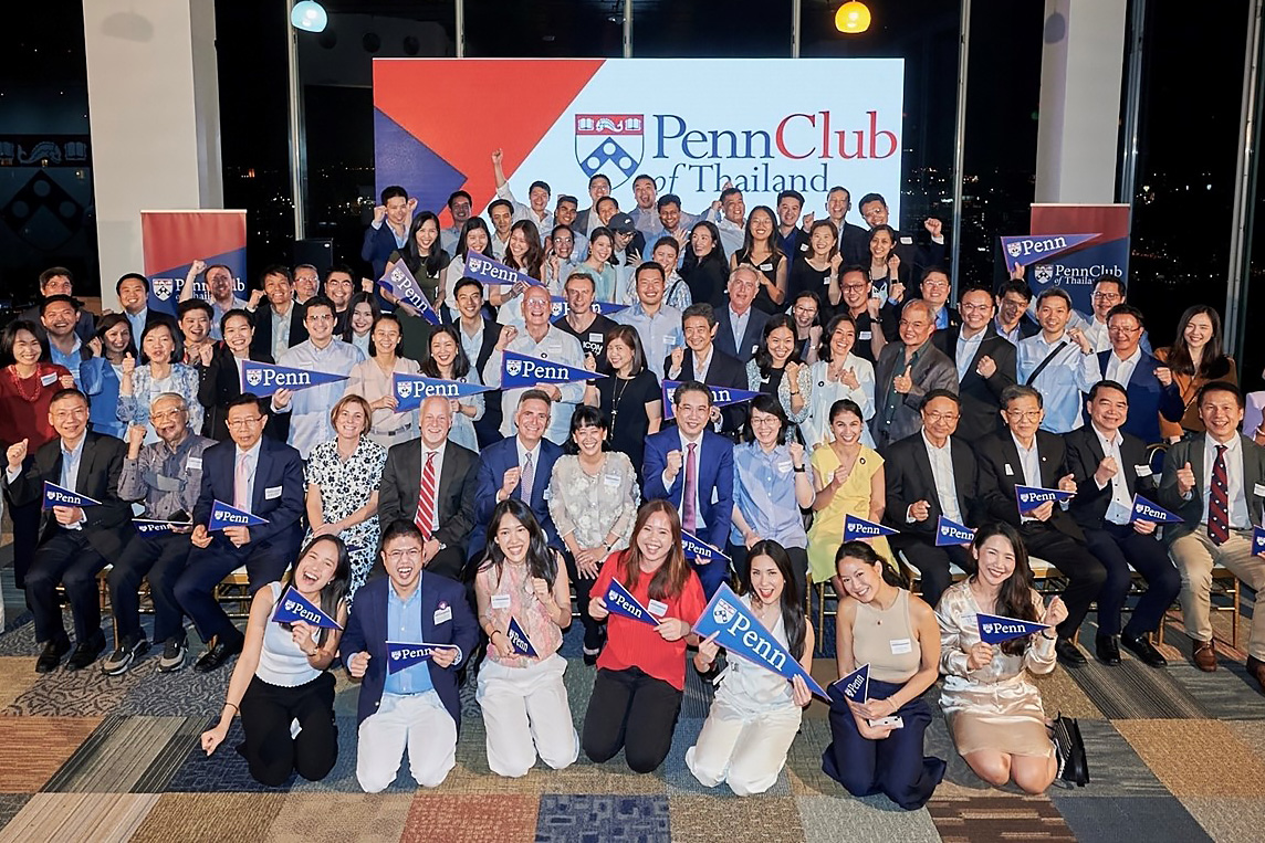 A vibrant gathering of the Penn Club of Thailand, featuring a diverse group of alumni from various ages and backgrounds, enthusiastically holding Penn banners and pennants.