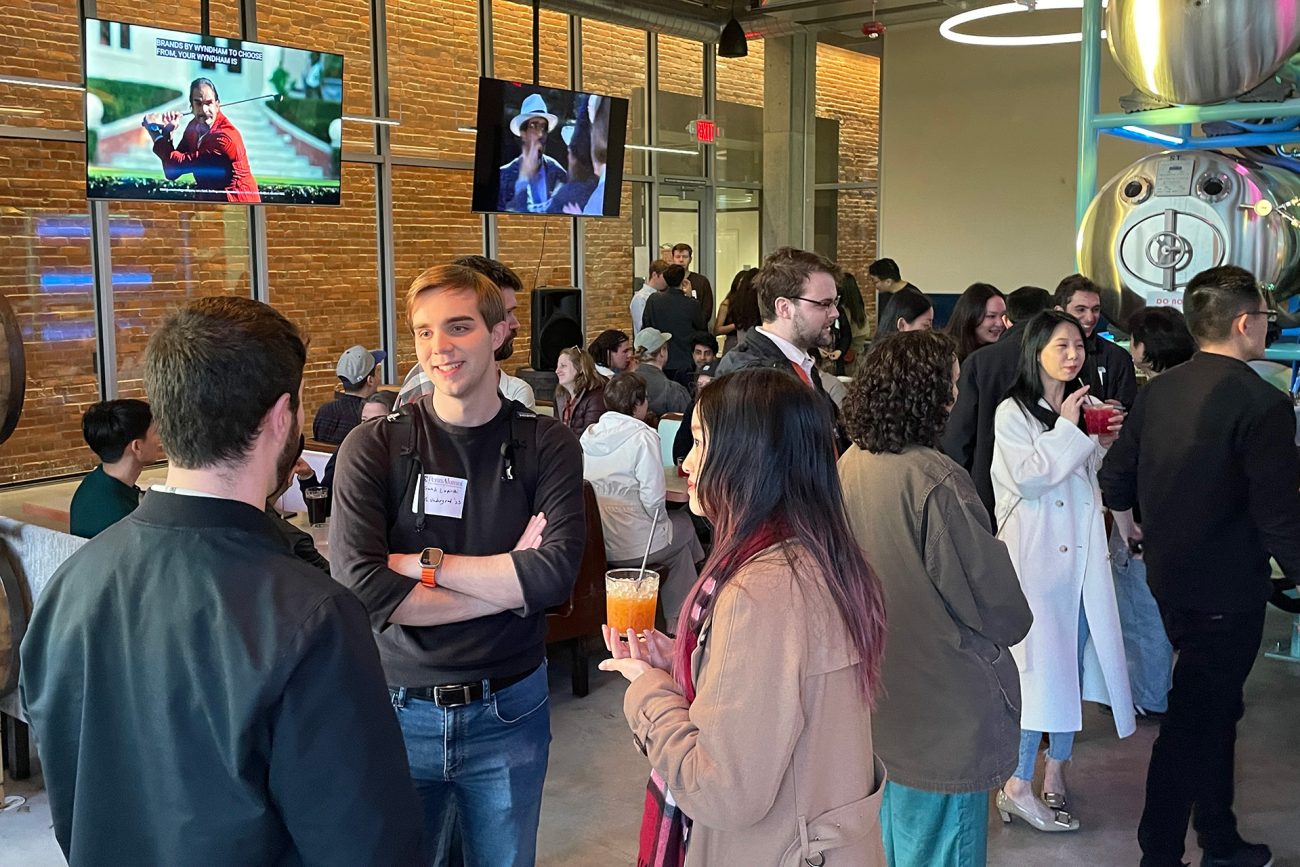 A lively networking event inside a modern brewery with rustic brick walls and brewing tanks visible. Diverse groups of young professionals engage in conversations.