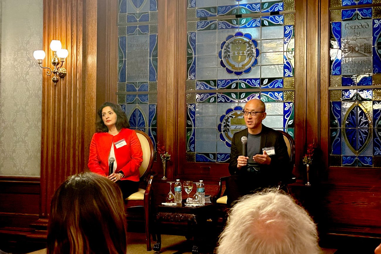 Two speakers, a woman in a red blazer and a man in a black sweater, are seated in front of an audience in a room with ornate blue stained glass windows and dark wooden paneling. The woman looks contemplative while the man speaks into a microphone, both addressing the attentive crowd gathered for the event.