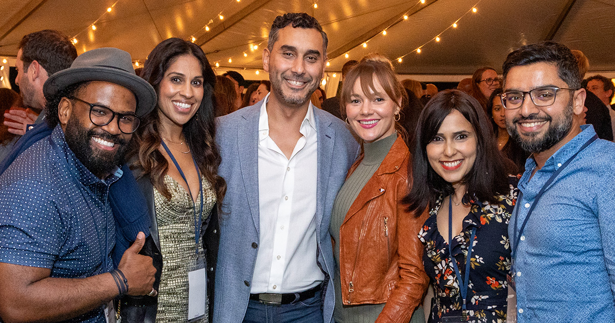 A vibrant group of six adults pose together at an outdoor event under a tent adorned with string lights. They are dressed in stylish casual and semi-formal attire, each wearing a smile, capturing a moment of joyful social interaction during the gathering.