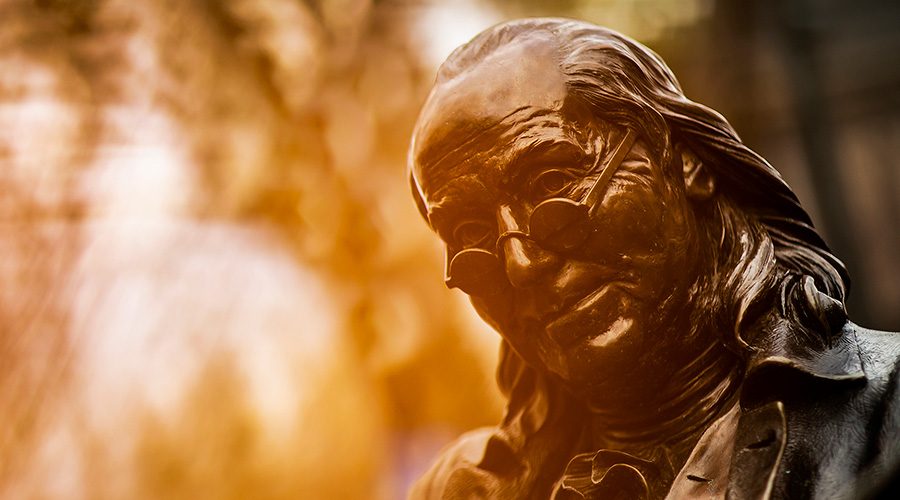 Close-up of a bronze statue of Benjamin Franklin, portrayed with a slight smile and wearing spectacles. The background is softly blurred, with warm golden and amber tones highlighting the contours of Franklin’s face and attire.