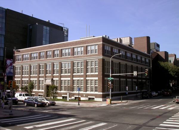 A wide-angle view of the Moore School Building, a large, traditional three-story school building located at the corner of 33rd and Walnut Streets on the University of Pennsylvania campus. The building features a red brick facade with long rows of white-framed windows.