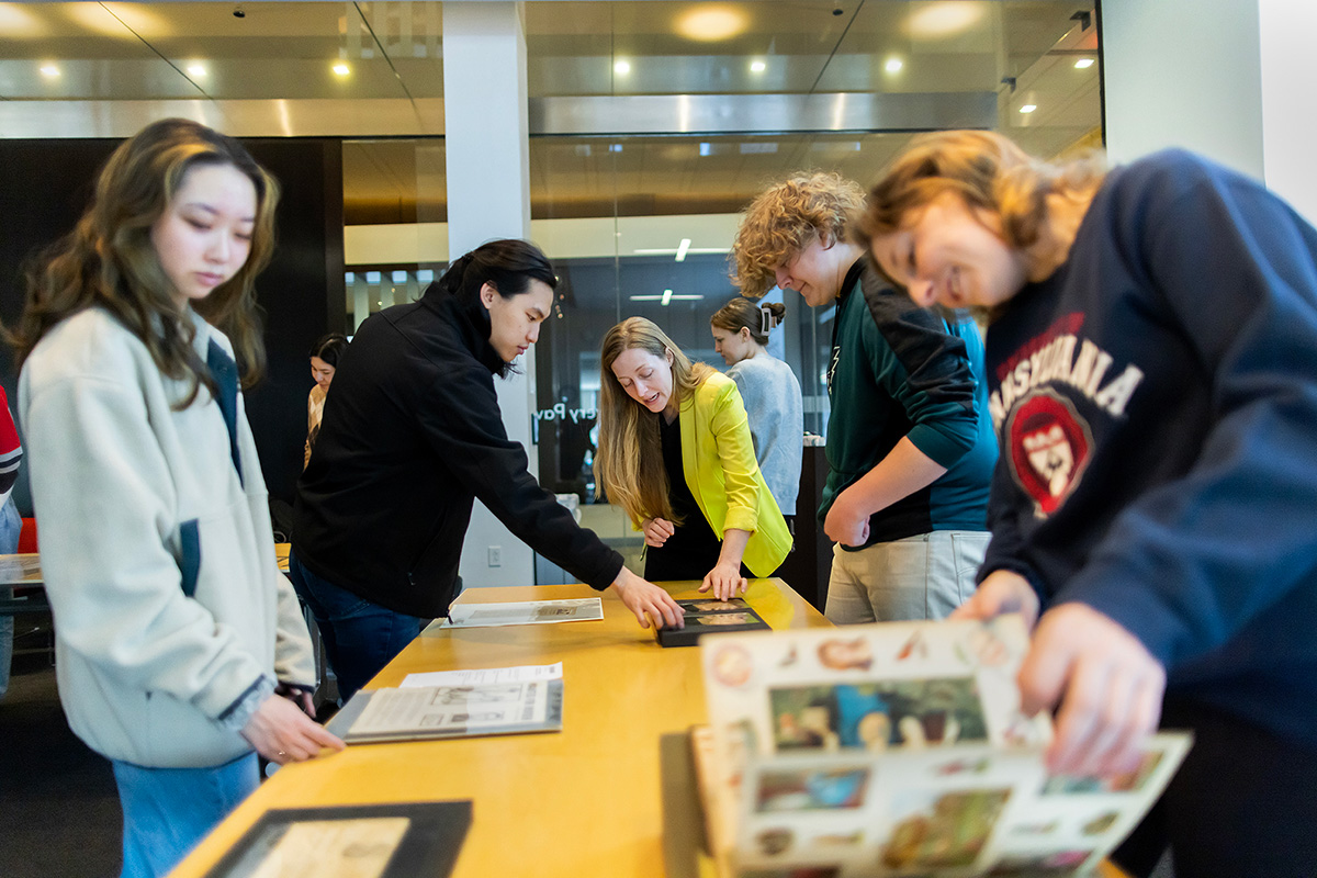 A group of students of diverse backgrounds engage in an interactive display at the University of Pennsylvania's Van Pelt-Dietrich Library Center. They are gathered around a long table, examining and discussing various printed materials laid out before them.