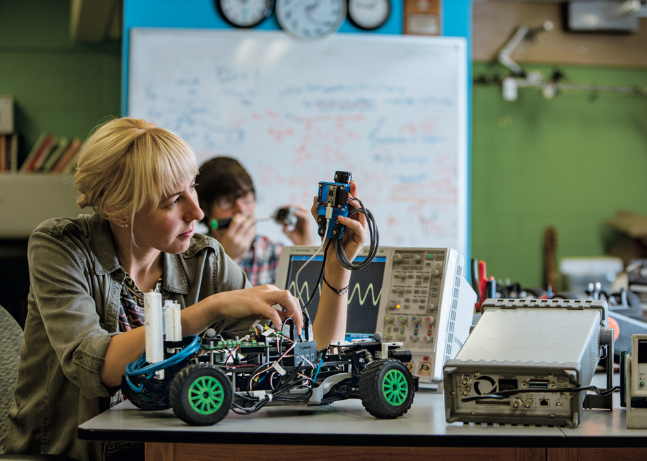 In a well-equipped robotics lab, a student with blonde hair is intently adjusting components on a sophisticated robotic vehicle. The environment is bustling with educational tools like oscilloscopes and other electronic testing equipment.