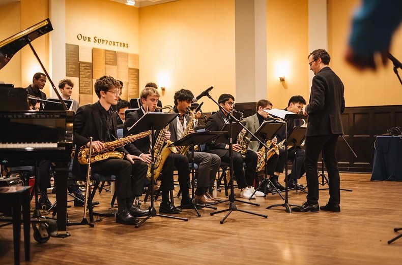A school jazz band performs in a well-lit room with walls adorned with 'Our Supporters' plaques. The young musicians, a diverse group of students, are seated in a semi-circle formation, attentively playing their instruments under the direction of a conductor.