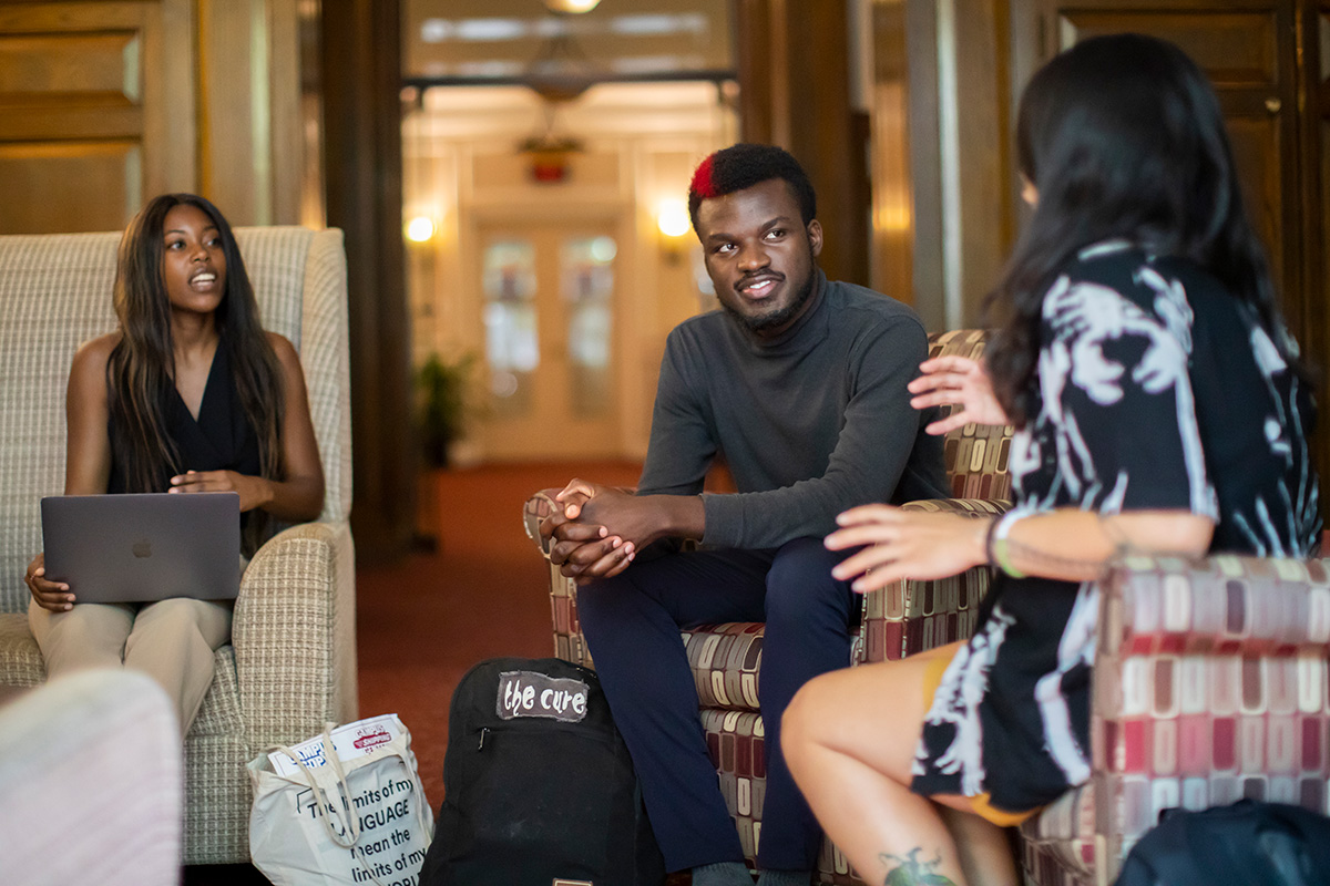 A candid scene in a warmly lit, vintage-style lobby where three students from the Social Justice Scholars Program engage in a lively discussion.