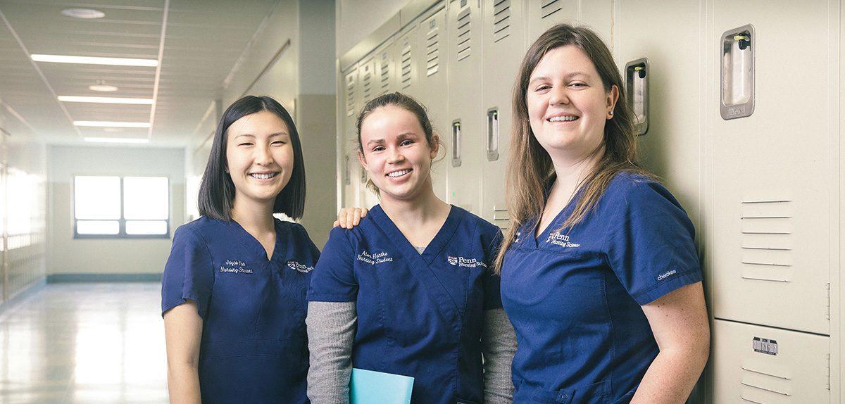Three Penn Nursing students in blue scrubs stand smiling in a school hallway. They are beside a row of gray lockers, each holding folders, suggesting a break between classes.