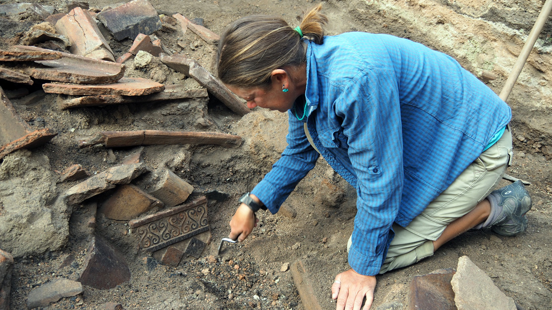 An archaeologist is intently excavating an ancient site. She carefully uses a small brush and trowel to uncover artifacts amidst a pile of broken terracotta pieces and dark soil.