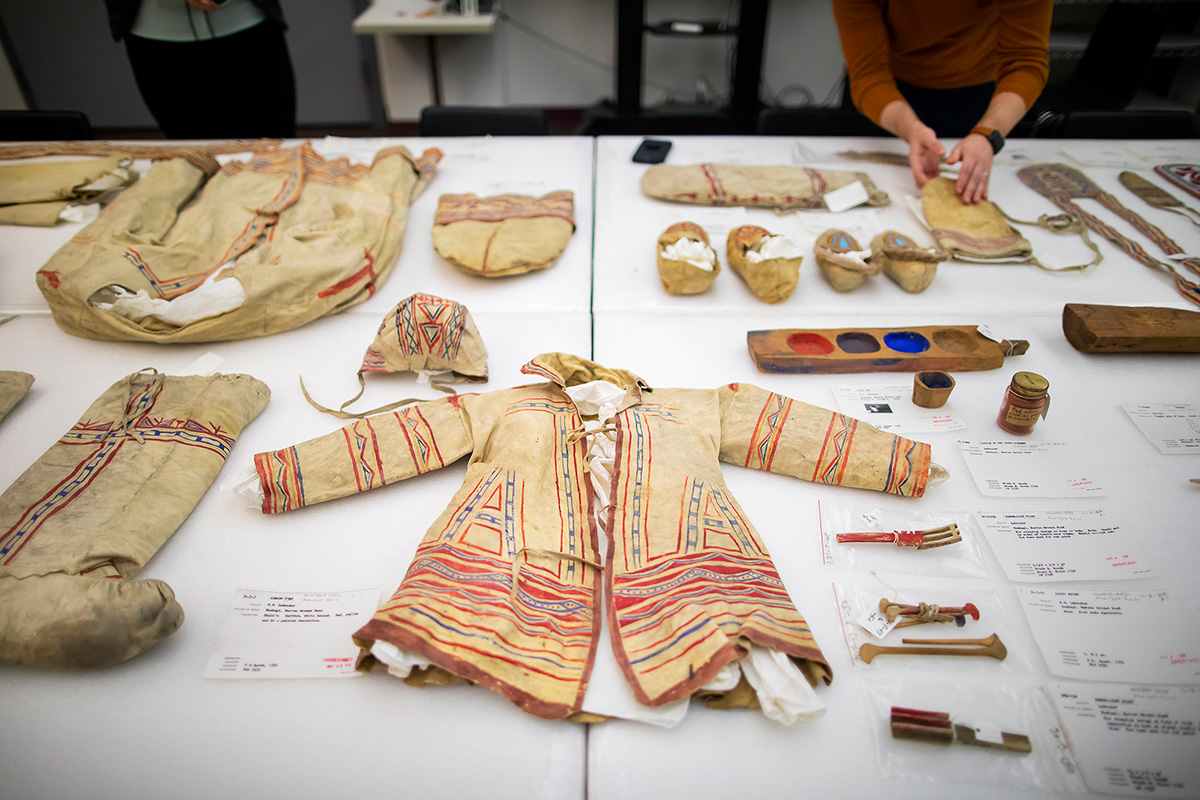 A collection of meticulously preserved ancient artifacts displayed on a white table within a museum setting. These objects include a small coat adorned with intricate red and orange geometric patterns, alongside smaller items like fabric shoes, painted wooden items, and small cosmetic containers. In the background, a person in an orange shirt is handling one of the objects.