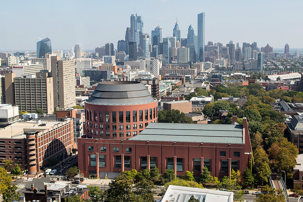 Aerial view of a bustling urban landscape in Philadelphia, showcasing a blend of modern skyscrapers and historic architecture. The foreground features Jon M. Huntsman Hall, a distinctive round-topped red building with a dark grey roof.