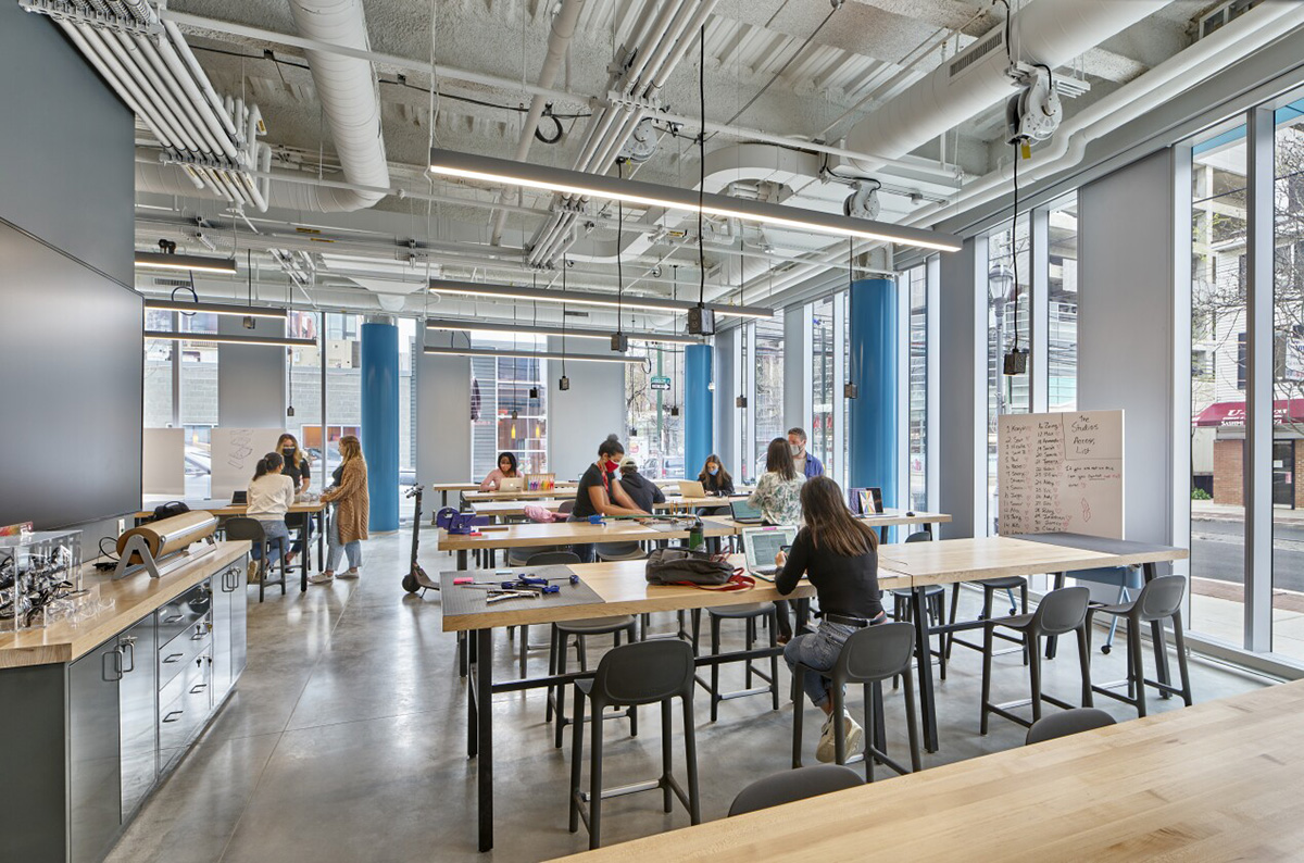 A workshop space inside Tangen Hall with students engaged in various activities. Several students are seated at long wooden tables equipped with laptops and other learning materials, collaborating in small groups.