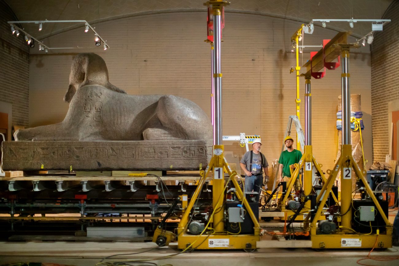 Two engineers stand beside the sphinx statue in the Penn Museum's lower Egyptian gallery. The granite statue is positioned on a complex moving platform, surrounded by mechanical lifts and yellow construction equipment used for its careful relocation.