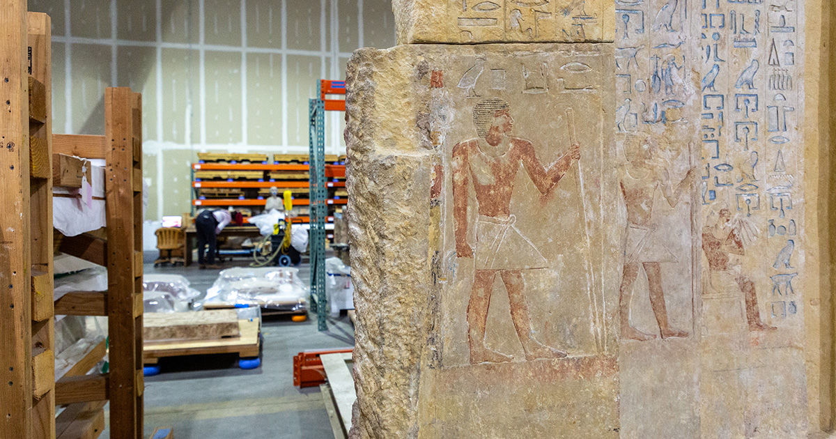 An ancient Egyptian relief, richly adorned with hieroglyphs and depictions of figures in traditional attire, is temporarily stored in a modern warehouse setting. The relief features a prominent figure in a standing pose, holding a staff, and is flanked by smaller detailed carvings. The background includes a visible storage area with wooden pallets and industrial shelves, filled with various items under protective covers.