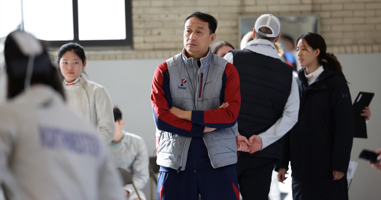 A focused middle-aged fencing coach wearing a gray and red team jacket with 'Fencing' embroidered on it, stands with his arms crossed in a gymnasium. He is observing a group of young athletes discussing strategy in the background.