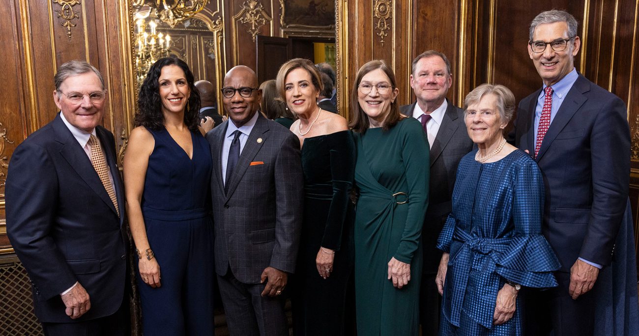 A group of eight individuals pose for a formal photo in an ornate room with rich wood paneling and golden chandeliers.