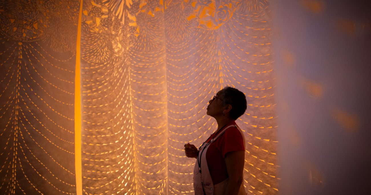 A woman, dressed in a red shirt and white overalls, stands mesmerized by an intricate wall art display featuring a complex pattern of golden spirals and floral motifs.