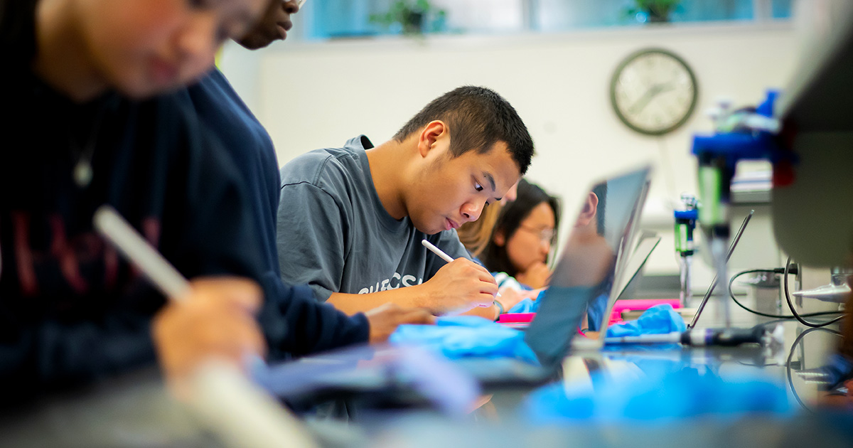 A focused young man in a gray sweatshirt is seen writing in a notebook in a classroom setting, surrounded by other students who are also engaged in their studies.