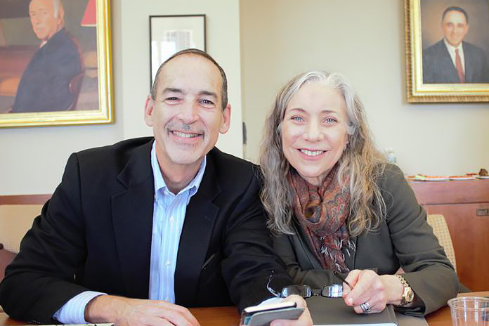 A smiling man and woman sitting at a table with portraits on the wall behind them.