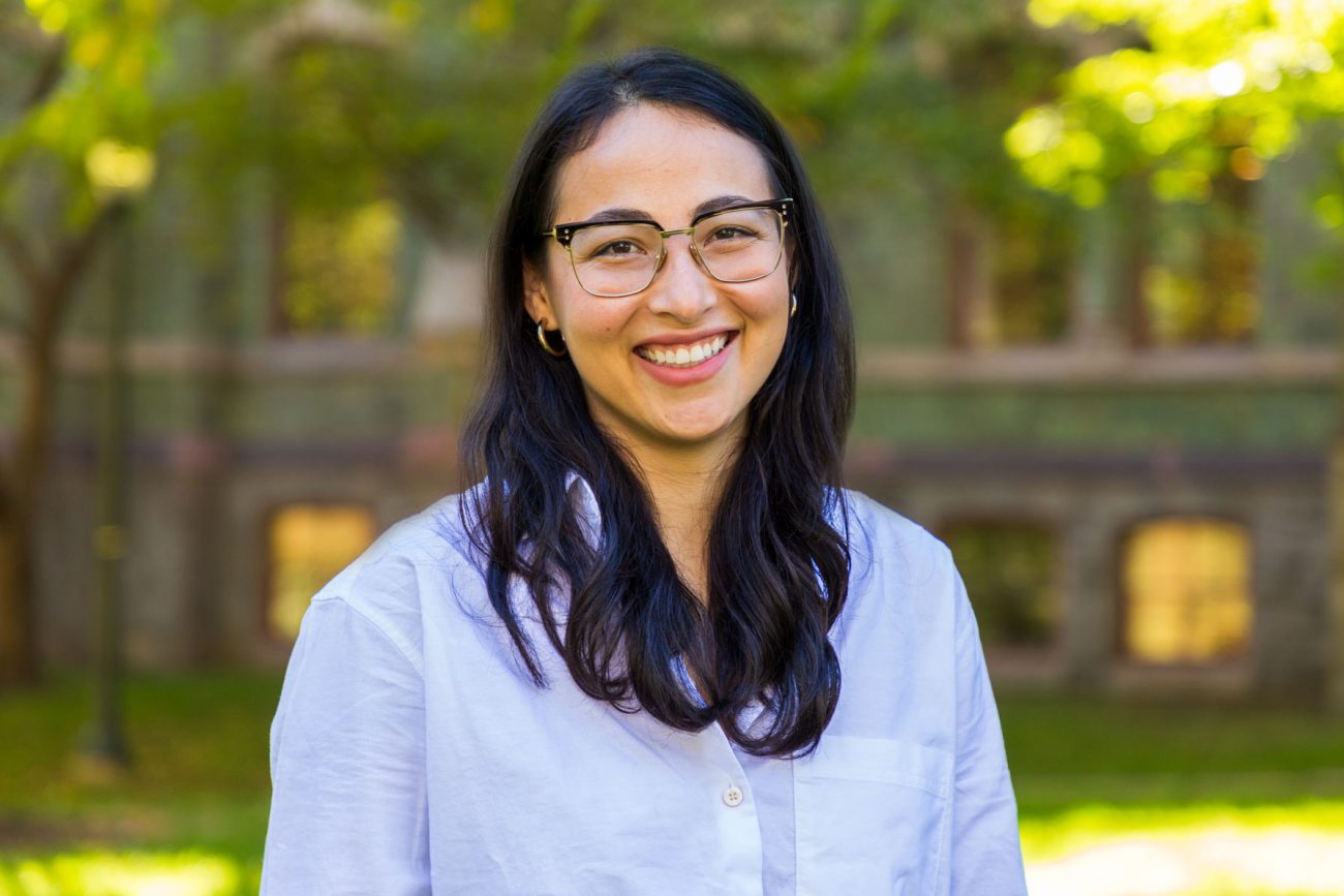 A young woman with glasses and long black hair smiles for the camera as she stands on College Green in front of College Hall.