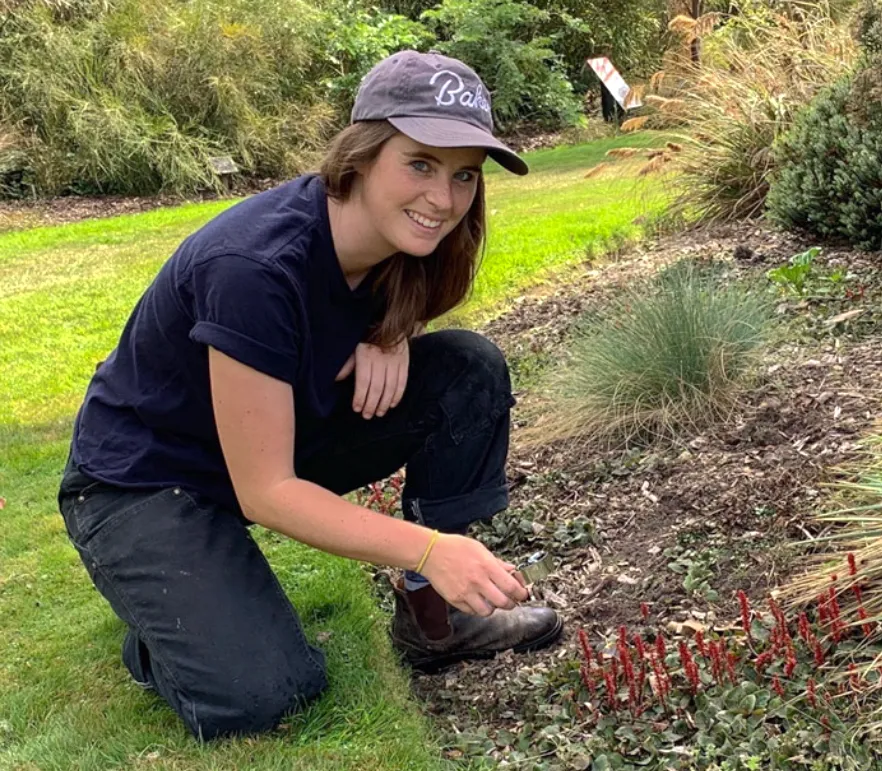 Kyra Matin crouches on the grounds of Morris Arboretum & Gardens, smiling while tending to red plants with a small gardening tool.