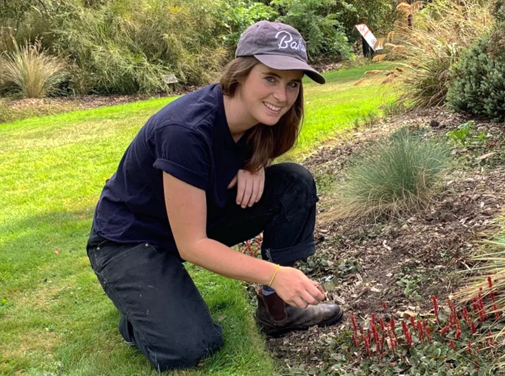 Kyra Matin crouches on the grounds of Morris Arboretum & Gardens, smiling while tending to red plants with a small gardening tool.