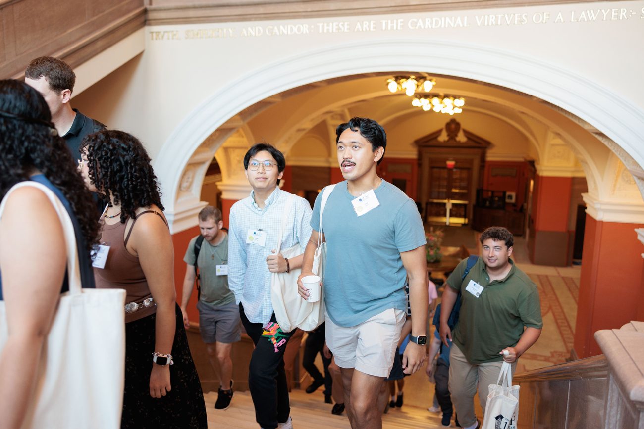 Students walking through an ornate university hallway, engaged in conversation during a Penn Carey Law event. The scene captures a moment of active engagement among a diverse group of attendees.