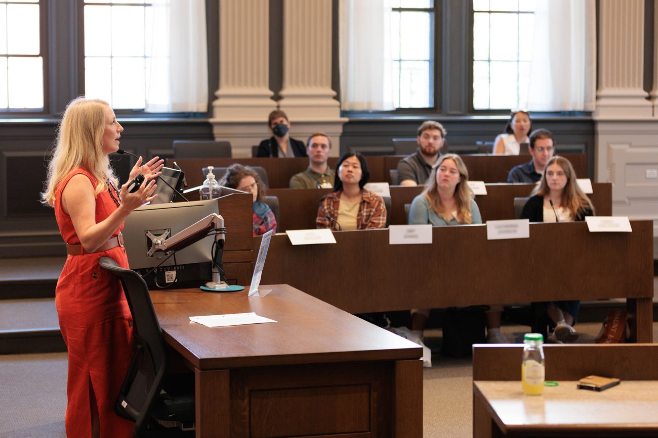 A focused classroom setting with a woman in a bright red dress presenting enthusiastically at the podium. The lecture hall is filled with attentive students seated at wooden desks, each with name placards, displaying a diverse group engaged in the lecture.