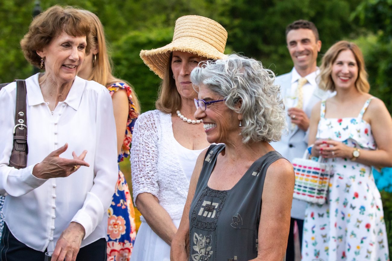 A group of cheerful guests at the Morris Arboretum & Gardens' annual Moonlight & Roses celebration, engaged in animated conversation. Jan Albaum, shown in the foreground, smiles broadly while others enjoy the moment. The setting is lush and green, enhancing the joyful, natural atmosphere of the gathering.