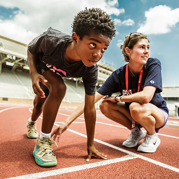 A young student athlete is shown in a starting position on the track at Franklin Field at the University of Pennsylvania, with an adult crouching next to him and looking onward.
