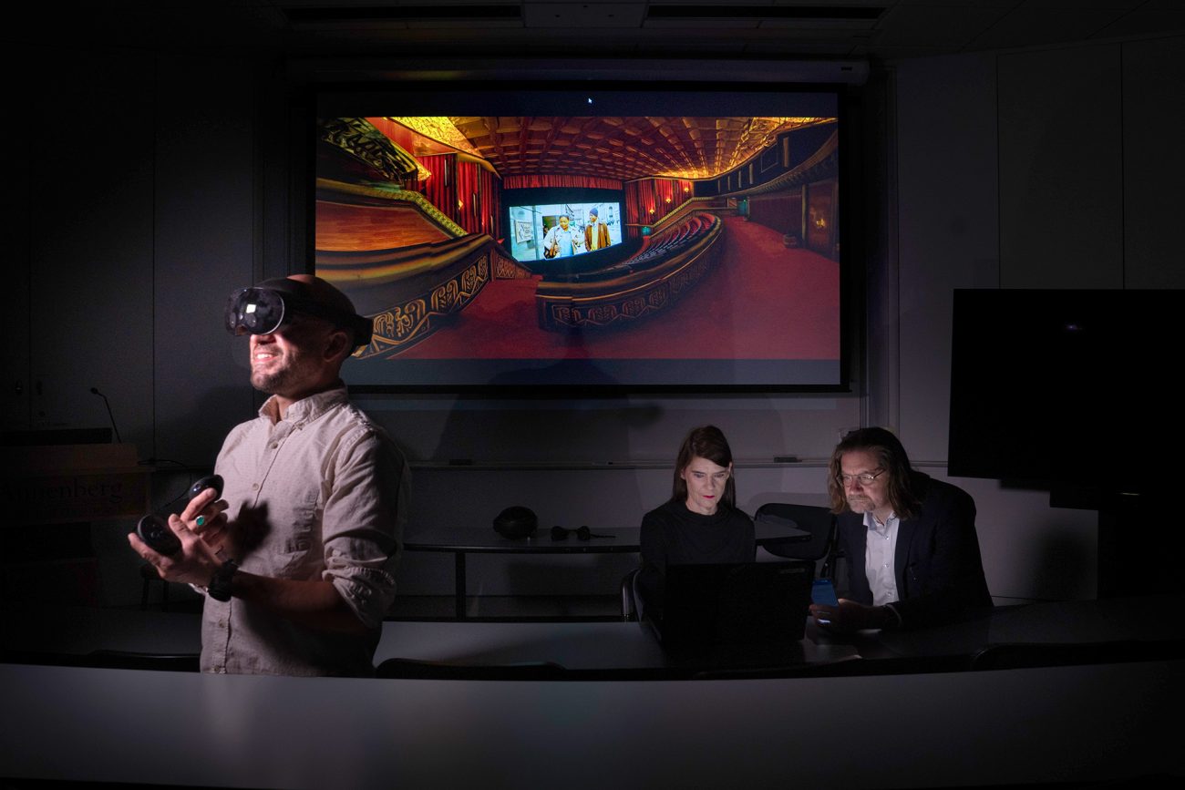 A person in a VR headset is actively engaged in a virtual reality demonstration in a dimly lit room. Behind them, two observers seated at a table watch the experience unfold on a large screen, which displays a colorful, ornate virtual environment. The scene is set in an educational context, highlighting the use of VR technology.