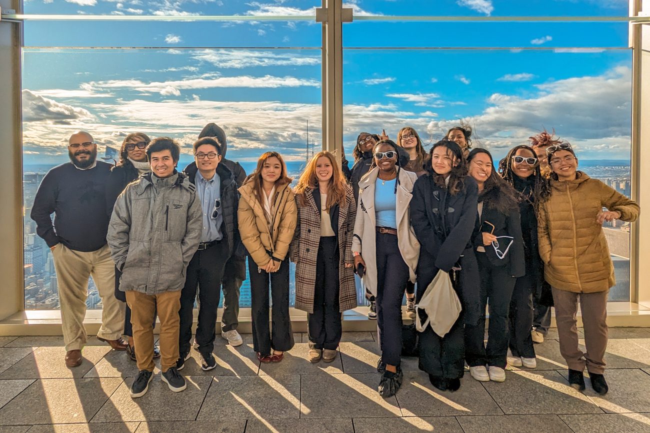 A group of students stand together in front of a window with the New York City skyline behind them