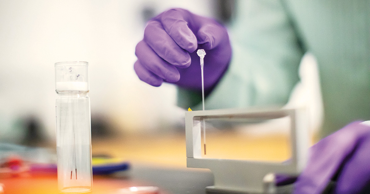 A close-up view of a laboratory setting showing a scientist's hand in a purple glove holding a pipette, extracting a small sample from a clear test tube. The background is softly blurred with hints of laboratory equipment and a bright, clean environment, emphasizing the precision and focus required in scientific research.