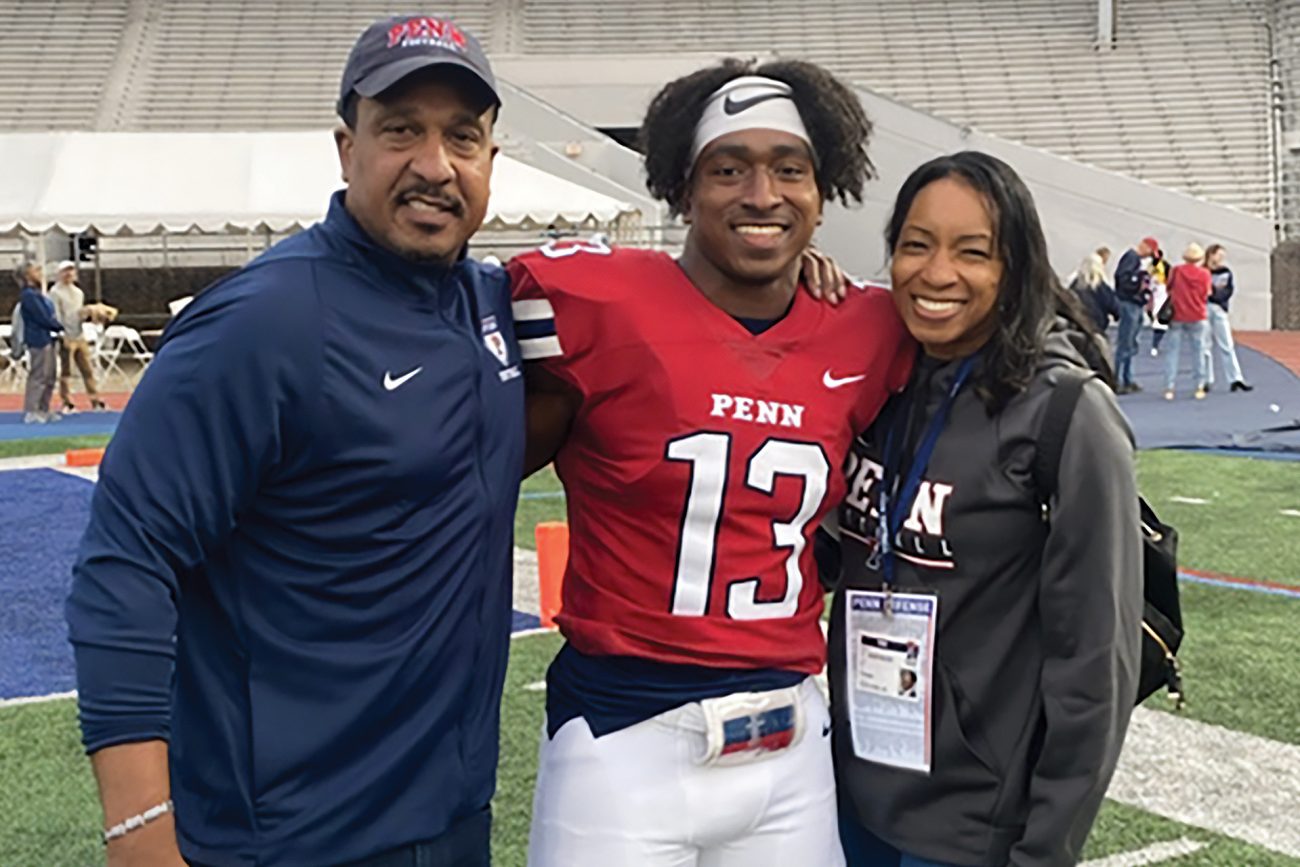 A joyful moment at a sports event with a college football player wearing a Penn jersey number 13, standing on the field with his proud parents. They are all smiling broadly, conveying a sense of family pride and achievement.