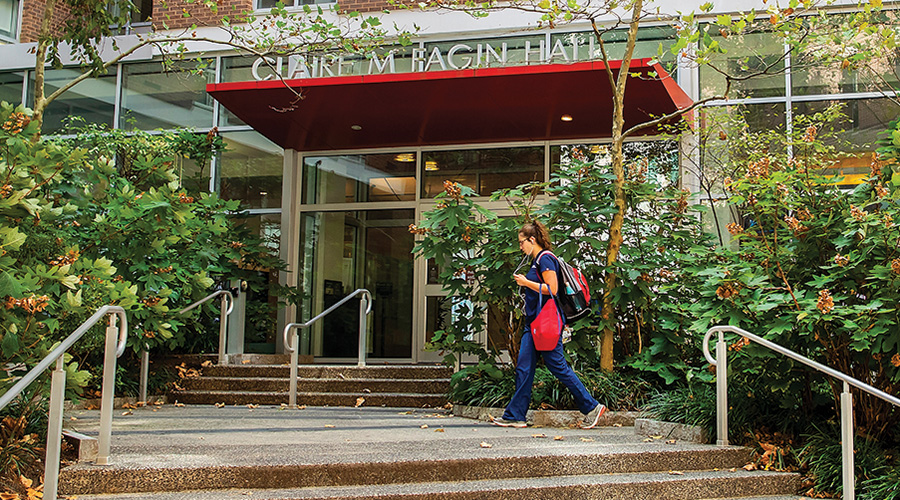 Outdoor scene at Claire M. Fagin Hall, where a student is ascending the steps towards the glass door entrance. The student is dressed in blue nursing scrubs with a red and black backpack. The building's entrance is surrounded by lush greenery and mature trees, enhancing the serene academic atmosphere.