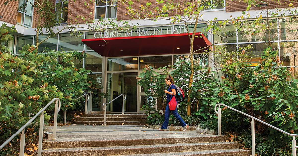Outdoor scene at Claire M. Fagin Hall, where a student is ascending the steps towards the glass door entrance. The student is dressed in blue nursing scrubs with a red and black backpack. The building's entrance is surrounded by lush greenery and mature trees, enhancing the serene academic atmosphere.